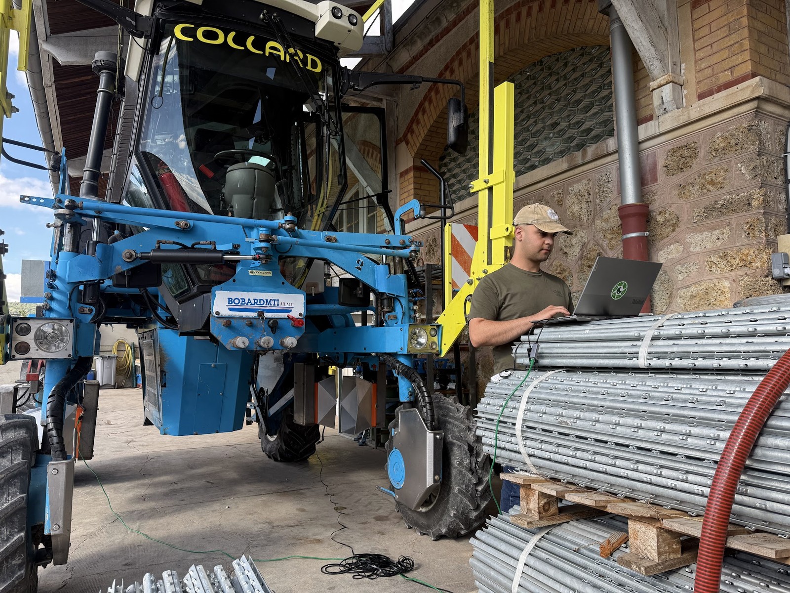 A Cropsy team member at a laptop commissioning newly fitted scanners on a blue Bobard straddle tractor in a French vineyard machinery hall