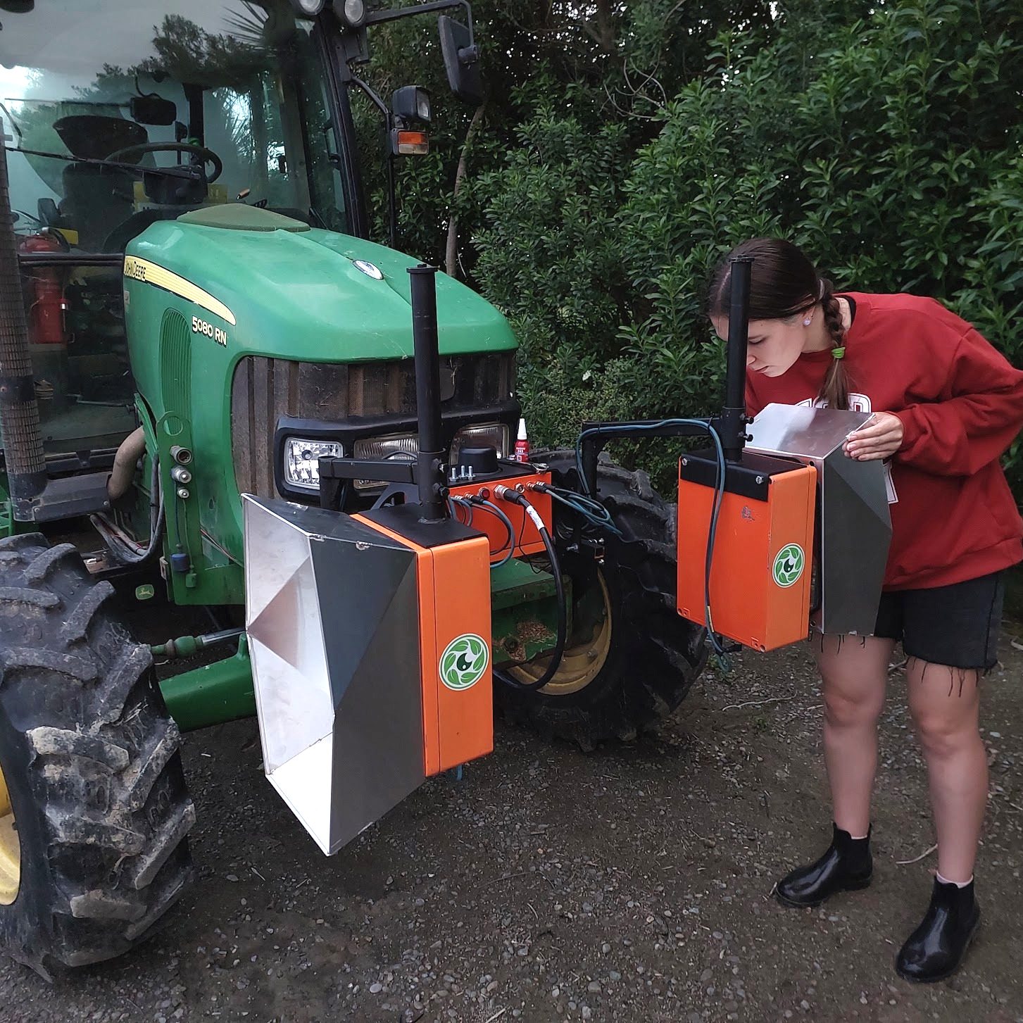 A green tractor fitted with two orange Cropsy scanner units and white calibration targets, a team member alongside taking notes