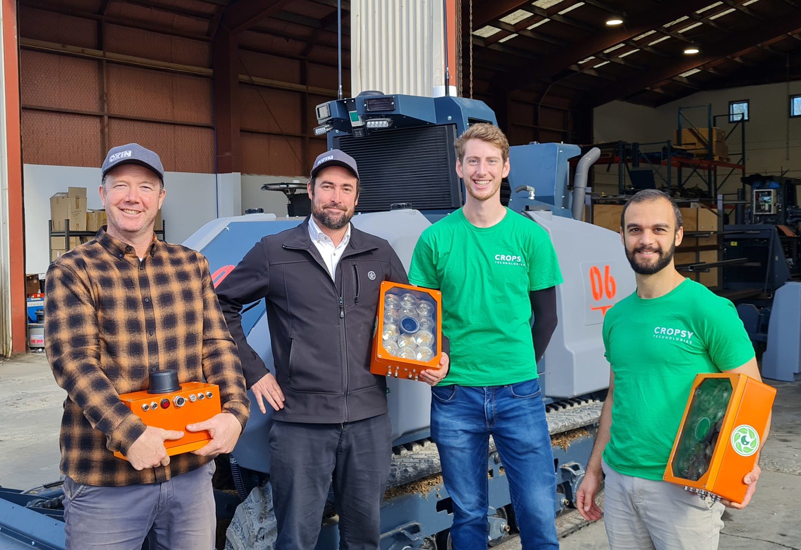 Four people in a machinery shed, each holding an orange Cropsy scanner module, in front of a vineyard machine